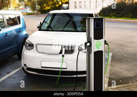Ein weißes Elektroauto lädt auf einem ausgewiesenen Parkplatz für Elektrofahrzeuge auf. Neben dem Fahrzeug ist eine Ladestation mit grünen Kabeln c zu sehen Stockfoto