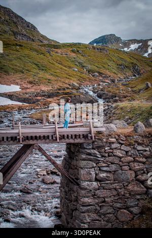 Norwegen. Ein Mädchen, das auf einer hölzernen Brücke über einen Bergfluss steht. Im Hintergrund sind mit Schnee und Moos bedeckte Berggipfel zu sehen. Stockfoto