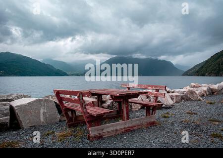 Ein Ruheplatz mit malerischer Aussicht. Ein Tisch und zwei Bänke stehen am Ufer mit Blick auf einen Fjord, Berge mit Wald und Nebel. Stockfoto
