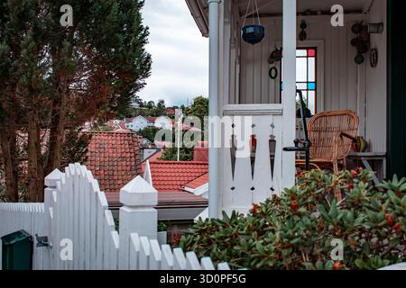 Eine bezaubernde Szene im Freien mit einem weißen Pflaumenzaun, einer Veranda mit einem Korbstuhl und einem Blick auf rot gekachelte Dächer und weiße Häuser im Hintergrund. Stockfoto