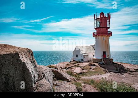 Ein malerischer Leuchtturm an einer felsigen Küste mit einem kleinen, weißen Gebäude, mit Blick auf das weite, ruhige Meer unter einem hellblauen Himmel. Stockfoto
