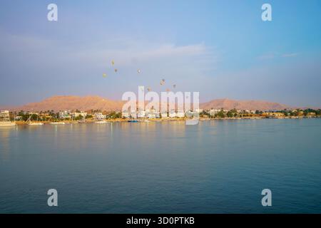 Farbenfrohe Heißluftballons steigen bei Sonnenaufgang über das Westufer von Luxor und die Wüstenberge auf, von den friedlichen Gewässern des Nils in Ägypten aus gesehen. Stockfoto