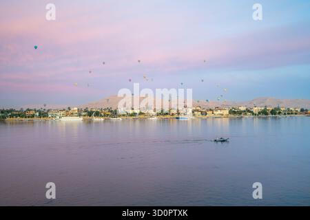 Ruhiger Blick auf die Dämmerung von der Bootstour auf den Nil mit farbenfrohen Heißluftballons, die im Frühjahr 2025 über Luxor West Bank, Ägypten, mit Wüstenhügeln und schwimmen Stockfoto
