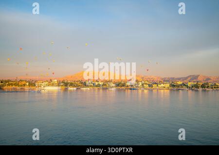 Ruhiger Blick auf die Dämmerung von der Bootstour auf den Nil mit farbenfrohen Heißluftballons, die im Frühjahr 2025 über Luxor West Bank, Ägypten, mit Wüstenhügeln und schwimmen Stockfoto
