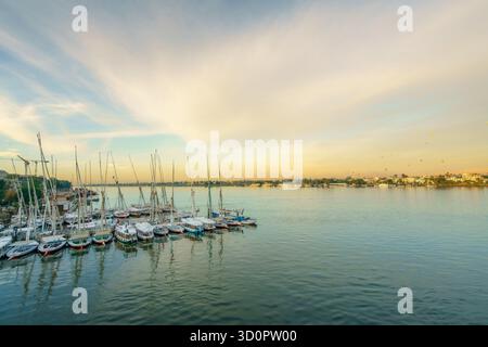 Traditionelle ägyptische Felucca-Segelboote liegen bei Sonnenaufgang auf dem ruhigen Nil in Luxor, Ägypten, mit schimmernden Wolken und dem sanften Glanz des Frühlingssonnenaufgangs Stockfoto
