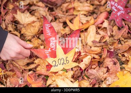Hand mit gelbem Ahornblatt, auf dem „Happy New Year 2026“ steht – Herbstgrußkonzept Stockfoto