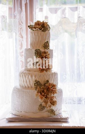 Eine atemberaubende, vierstufige weiße Hochzeitstorte mit strukturiertem Froster, goldfarbenen tropischen Blättern und zarten orangen Blumendetails. Perfekt Stockfoto
