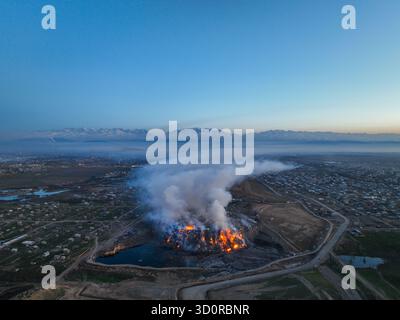Panoramablick auf eine große Mülldeponie, die mit hellen Flammen und einer riesigen Rauchwolke über der nahe gelegenen Stadt und den schneebedeckten Bergen brennt Stockfoto