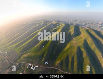 Blick aus der Vogelperspektive auf sonnendurchflutete, grüne Ausläufer und Terrassenfelder, die bei einem trüben Sonnenuntergang zu kleinen landwirtschaftlichen Gebäuden führen Stockfoto
