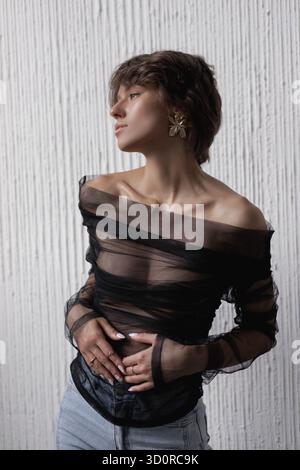 Die Frau steht selbstbewusst und trägt ein transparentes schwarzes Oberteil, das ihre Eleganz unterstreicht. Sie posiert vor einer strukturierten weißen Wand und zeigt ihr Outfit und einen Stockfoto