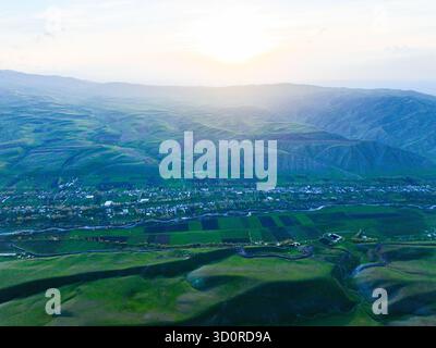 Weitläufiger Blick aus der Luft auf ein weites Tal mit Fluss und Dorf, umgeben von massiven, sanften grünen Hügeln unter einem hellen, trüben Sonnenuntergang Stockfoto