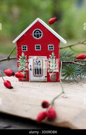 Bezauberndes rotes Miniaturhaus mit handgemalten Details, eingebettet zwischen Hagebutten auf einer rustikalen Holzoberfläche mit weichem grünem Hintergrund. Stockfoto