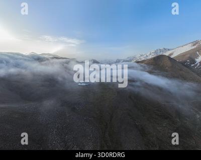 Weite Sicht aus der Luft auf einen hoch gelegenen, möglicherweise gefrorenen See, eingebettet in felsiges Gelände, mit Nebel oder Nebel, der dramatisch um die Hänge wirbelt Stockfoto