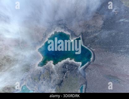 Abstrakte Ansicht von oben nach unten auf einen kleinen, tiefblauen Gletschersee mit unregelmäßiger Form, teilweise durch Wolken oder Nebel verdeckt und von Kies umgeben Stockfoto