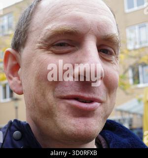 Ein Mann mittleren Alters mit kurzen Haaren ist draußen und trägt eine dunkle Jacke. Er erzählt eine Geschichte und lächelt ironisch Stockfoto