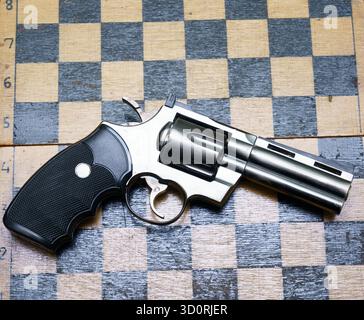Ein Revolver aus glänzendem Metall wird auf einem Schachbrett aus dunklen und hellen Holzquadraten platziert und sorgt so für einen markanten Kontrast. Stockfoto