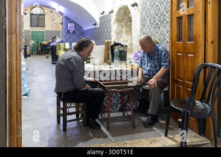 Jerusalem, Israel, 23. Oktober 2025, sitzen zwei Männer mittleren Alters auf kleinen Holzhockern und spielen Backgammon in einem traditionellen, gekachelten Raum im alten Cit Stockfoto