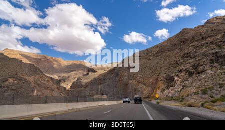 Zwei Fahrzeuge fahren auf einer Autobahn durch einen felsigen Bergpass unter blauem Himmel mit verstreuten Wolken. Stockfoto
