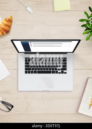 Vertikale Draufsicht des Laptops auf Holzschreibtisch mit Kaffee, Notebook, Croissant und Pflanze, symbolisiert digitale Arbeit und moderne Produktivität Stockfoto