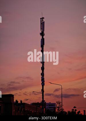 Ein Handymast vor der Kulisse eines pulsierenden Sonnenuntergangs und Stadtgebäude in warmen Abendtönen Stockfoto