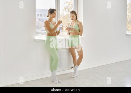Zwei Frauen in passenden grünen Workout-Outfits machen während eines Fitness-Stretchkurses in einem hellen Studio eine Pause. Sie reden und hydratisieren am Fenster, en Stockfoto