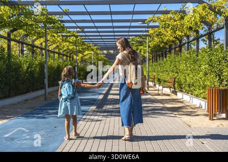 Mutter und Tochter gehen auf einem urbanen Pfad, halten Hände und tragen Rucksäcke an einem sonnigen Tag. Dieses Bild repräsentiert Themen der Familie, der Bildung Stockfoto