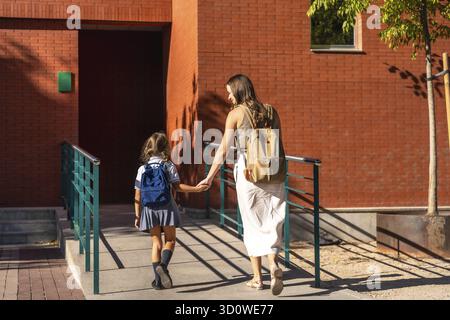 Mutter und Tochter gehen Hand in Hand mit Rucksäcken bergauf, kehren zur Schulbildung zurück, unterstützen und bereiten sich auf ein neues Schuljahr an einem sonnigen Tag vor Stockfoto