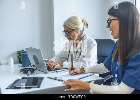 Zwei medizinische Fachkräfte, ein leitender Arzt, der auf einem Laptop tippt, und eine jüngere Krankenschwester, die Notizen auf einem Klemmbrett macht, arbeiten in einem modernen Gesundheitswagen zusammen Stockfoto
