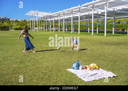 Mutter und Tochter spielen Fußball in einem sonnigen grünen Park, genießen ein Picknick mit Decke und Snacks, Bonding, aktive Erholung und Sommerspaß für die Familie Stockfoto