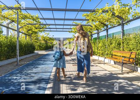 Mutter und Tochter gehen Hand in Hand auf einem urbanen Parkweg mit Rucksäcken, gehen auf eine sonnige Tagesschule, Führung und einen Neuanfang Stockfoto