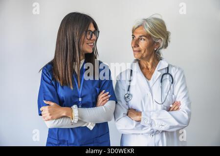 Zwei selbstbewusste Ärztinnen, eine jüngere und eine ältere, stehen mit gekreuzten Armen vor weißem Hintergrund, repräsentieren Teamarbeit und Gesundheitsfürsorge p Stockfoto