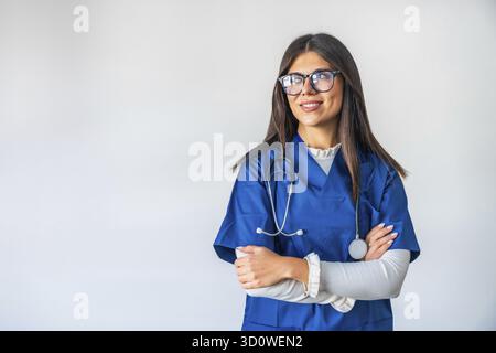Junge Frau, die als professioneller Gesundheitsdienstleister arbeitet, selbstbewusst mit gekreuzten Armen steht, zur Seite blickt und lächelt, Fachkompetenz verkörpert und Stockfoto
