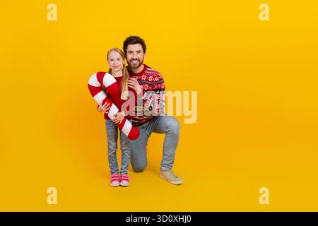 Fröhlicher Vater und Tochter posieren mit übergroßem Zuckerrohr auf gelbem Hintergrund in festlicher Kleidung Stockfoto