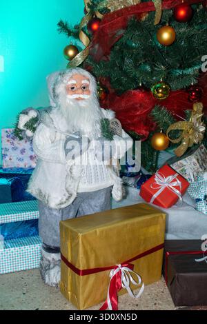 Ein Weihnachtsmann steht neben einem Weihnachtsbaum, der mit hellen Ornamenten verziert ist, umgeben von gewickelten Geschenken und bunten Lichtern. Stockfoto