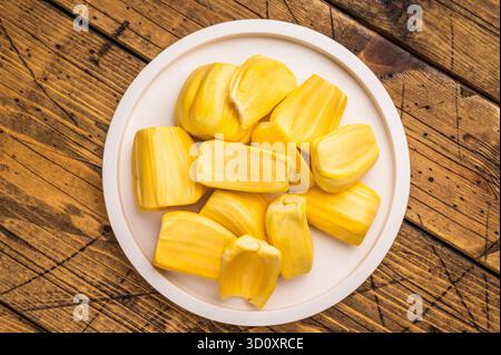 Süße, goldgelbe Jackfruchskapseln (Zwiebeln), serviert auf einem weißen Teller auf einem rustikalen Holztisch. Frischer, tropischer Obstsnack Stockfoto