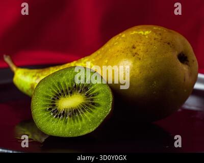 Frisches grünes Kiwi, das neben einer ganzen Reifen Birne auf einem dunklen Teller halbiert wird Stockfoto