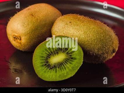 Frische Kiwi-Früchte, einschließlich einer in der Mitte geschnittenen Nahaufnahme - reife Kiwi auf dunklem Teller Stockfoto