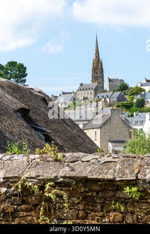 Ein bezauberndes strohgedecktes Cottage befindet sich neben einer verwitterten Mauer, mit dem malerischen Dorf Pont-Croix in der Bretagne und einem hohen Kirchturm im Hintergrund. Heller Himmel, üppiges Grün. Stockfoto