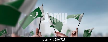 Pakistanische Flaggen in den Händen vor einem verschwommenen urbanen Hintergrund. Die Fotoillustration wurde ohne künstliche Intelligenz erstellt, nur mit Photoshop. Stockfoto