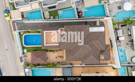 Blick von oben auf eine Wohnvilla mit einem komplexen, braun gekachelten Dach, rechteckigem Swimmingpool, überdachter Pergola, Dachterrasse und gepflastertem Ya Stockfoto