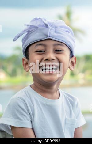 Nahporträt eines balinesischen Kindes, das am Galungan Day fröhlich lacht, im weißen zeremoniellen „Udeng“-Stirnband mit verschwommenem Reisfeld-Hintergrund. Stockfoto