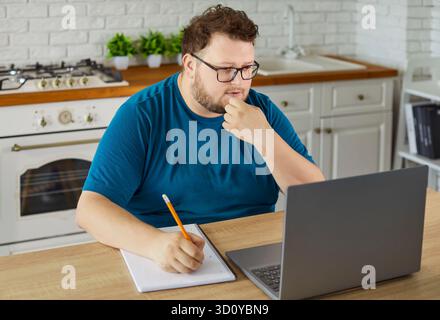 Mann in Übergröße, der online zu Hause in der Küche mit Laptop, Notebook und Bleistift studiert Stockfoto