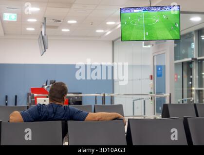 Ein Mann sitzt in einem ruhigen Wartebereich am Flughafenterminal und sieht sich ein Fußballspiel auf dem Fernseher an Stockfoto