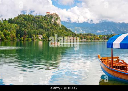 Ein traditionelles hölzernes Pletna-Boot entlang des Seeufers mit der mittelalterlichen Klippe und der Burg auf einem Hügel auf dem Bleder See, in Bleder Slowenien, Julischen Alpen. Stockfoto