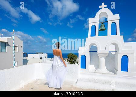 Eine attraktive brünette Frau in einem langen weißen Kleid überblickt das Meer von einem weißen Glockenturm in der weiß getünchten Stadt Oia, Santorin Griechenland Stockfoto