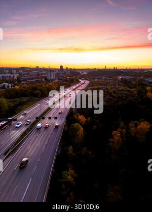 Blick aus der Vogelperspektive auf die Autobahn mit sechs Spuren, die zu einer urbanen Stadtlandschaft mit farbenfrohem Himmel bei Sonnenuntergang führen Stockfoto