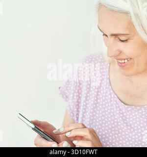 Ältere weiße Frau mit Smartphone in Polka Dot-Shirt lächelt auf Touchscreen-Display. Stockfoto