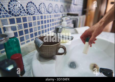 Eine Badezimmerszene zeigt einen Keramikbecher mit einem Teebeutel, einem grünen Seifenspender und einer Hand, die zum Waschbecken reicht, auf blauen Mosaikfliesen. Stockfoto