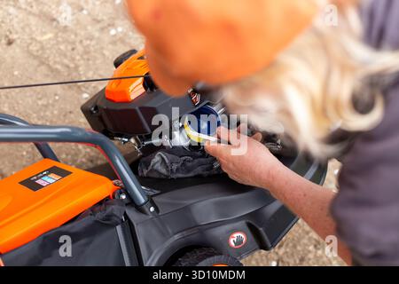Ein Mann überprüft den Ölstand im Motor eines Rasenmähers mit einem Messstab, führt einen Trichter ein und bereitet das Nachfüllen von Öl vor. Stockfoto