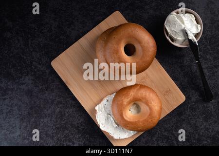 Ein hoher Blick auf zwei Bagels mit Frischkäse auf einem hölzernen Schneidebrett. Die Bagels werden in zwei Hälften geschnitten und der Frischkäse darüber verteilt Stockfoto